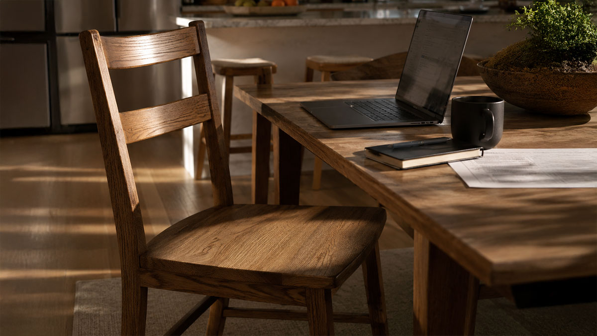 An empty solid wood dining chair pulled up to a kitchen table in late afternoon light, with an open laptop, a notebook and a mug on the table.