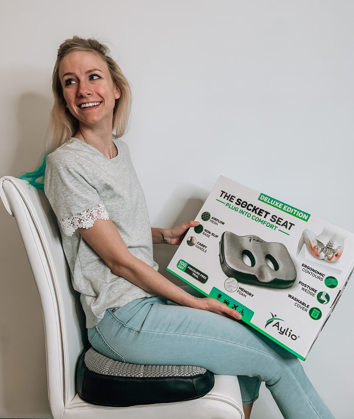 A smiling customer sits on her Socket Seat on a white chair, holding the retail box on her lap.